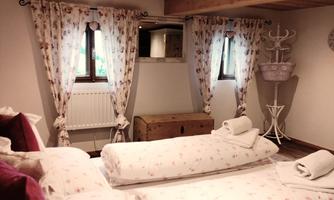 A cozy bedroom with a wooden ceiling and floral-patterned curtains. The bed is adorned with pillows and blankets, and a wooden wardrobe is visible in the background.