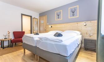 A cozy bedroom with two beds and modern wooden elements. The room features a red armchair and decorative pictures on the wall.