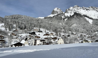 A picturesque winter village with snow-covered houses and an impressive mountain landscape. The trees are covered in snow, and the sky is clear and blue.