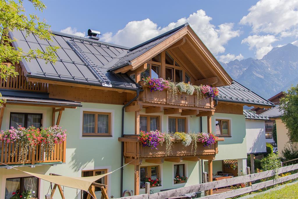 A charming house with a wooden balcony, adorned with colorful flowers. In the background, the mountains and a blue sky can be seen.
