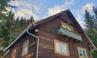A cozy wooden house in the forest with a balcony and green window frames. The sky is blue with some clouds.