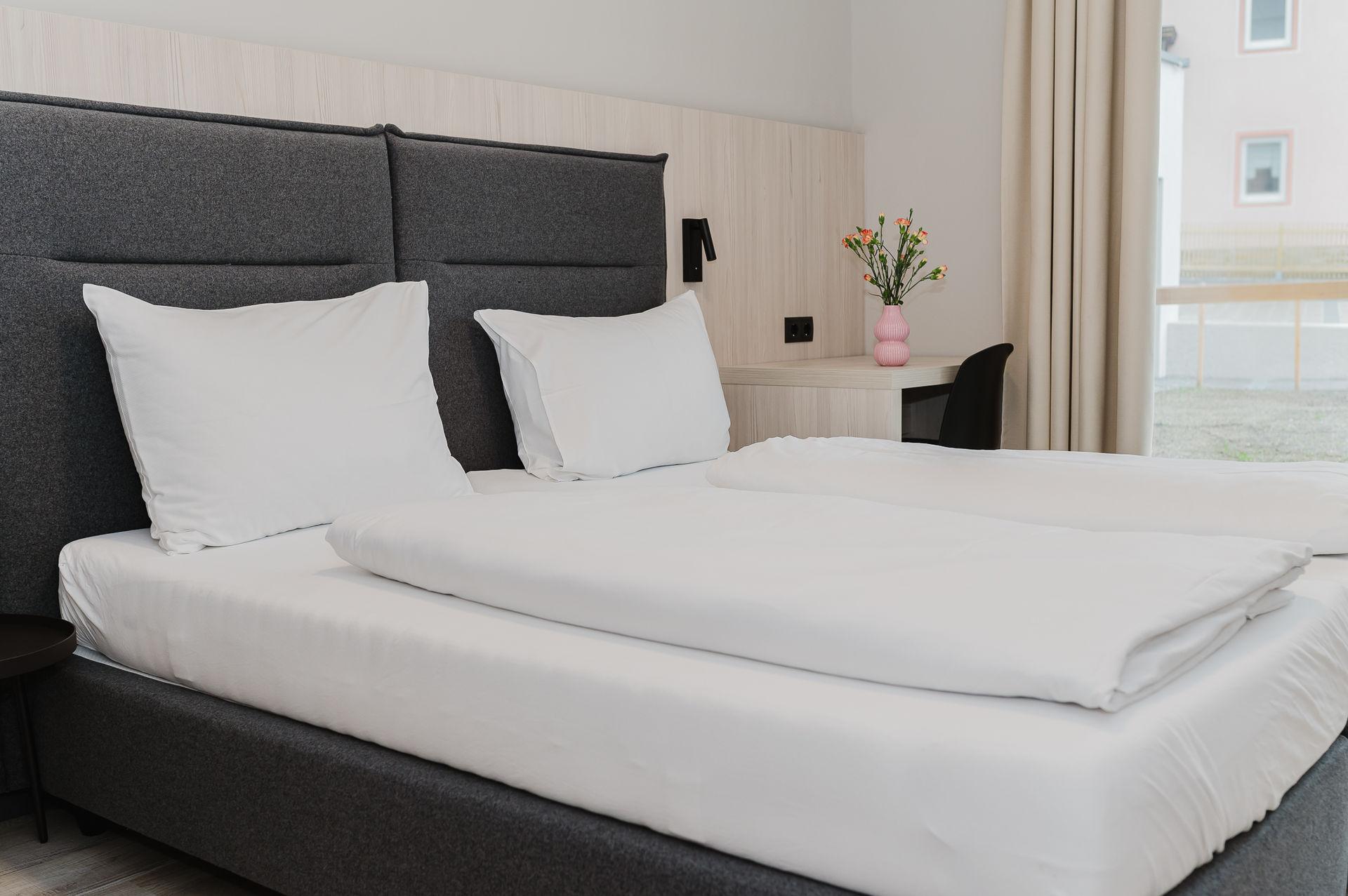 A modern hotel room with a double bed and white bedspreads. There is a desk and a small bouquet of flowers on the table.