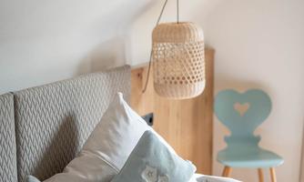 A cozy bedroom with a stylish bed and decorative pillows. A woven lampshade and a blue chair with a heart design add charm to the space.