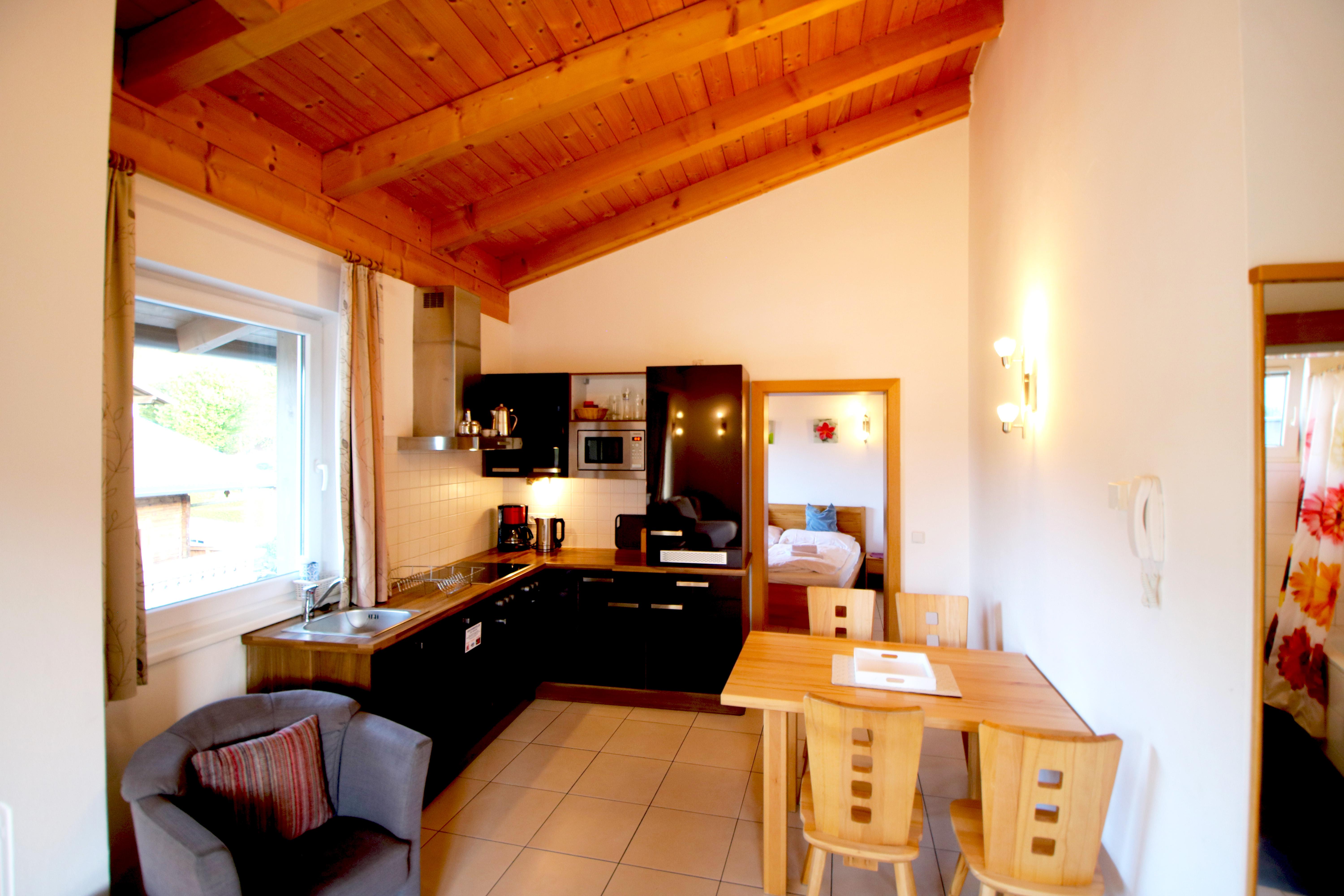 A modern kitchen with a wooden ceiling and open living area. There is a dining area with a table and chairs as well as a cozy armchair.