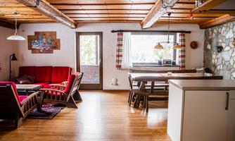 A cozy living space with wooden ceilings and warm colors. There is a seating area with red furniture and a dining table near a large window front.