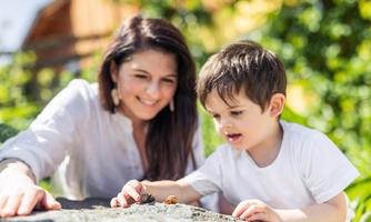 A cheerful child is playing with snails on a stone while an adult watches smilingly. The scene takes place outdoors in sunlight.