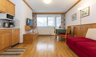A cozy apartment with wooden shelves and a red sofa. The room has plenty of daylight and a dining area with a table and chairs.