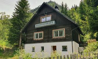 A traditional wooden house surrounded by trees and a green landscape. It has a balcony and a white base.