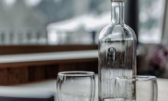 An elegant glass bottle stands on a wooden chair, accompanied by two clear glasses. Blurry mountains are visible in the background.