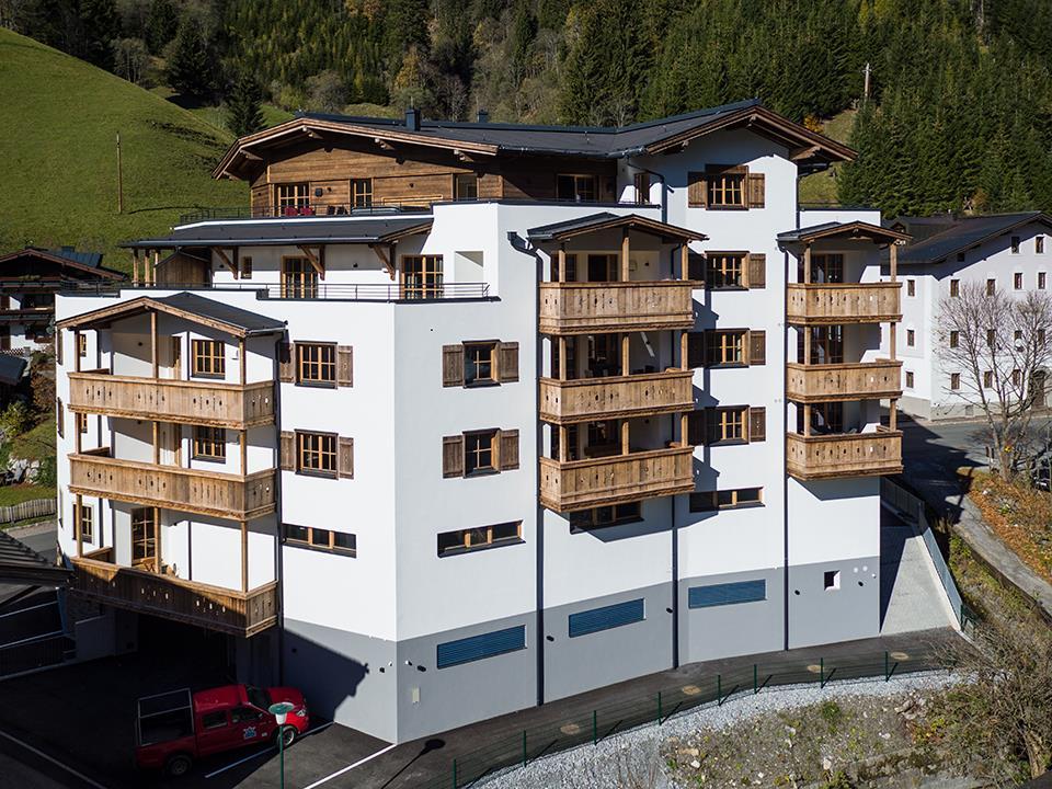 A modern building with wooden cladding and several balconies. It is located in a mountainous area, surrounded by green meadows.
