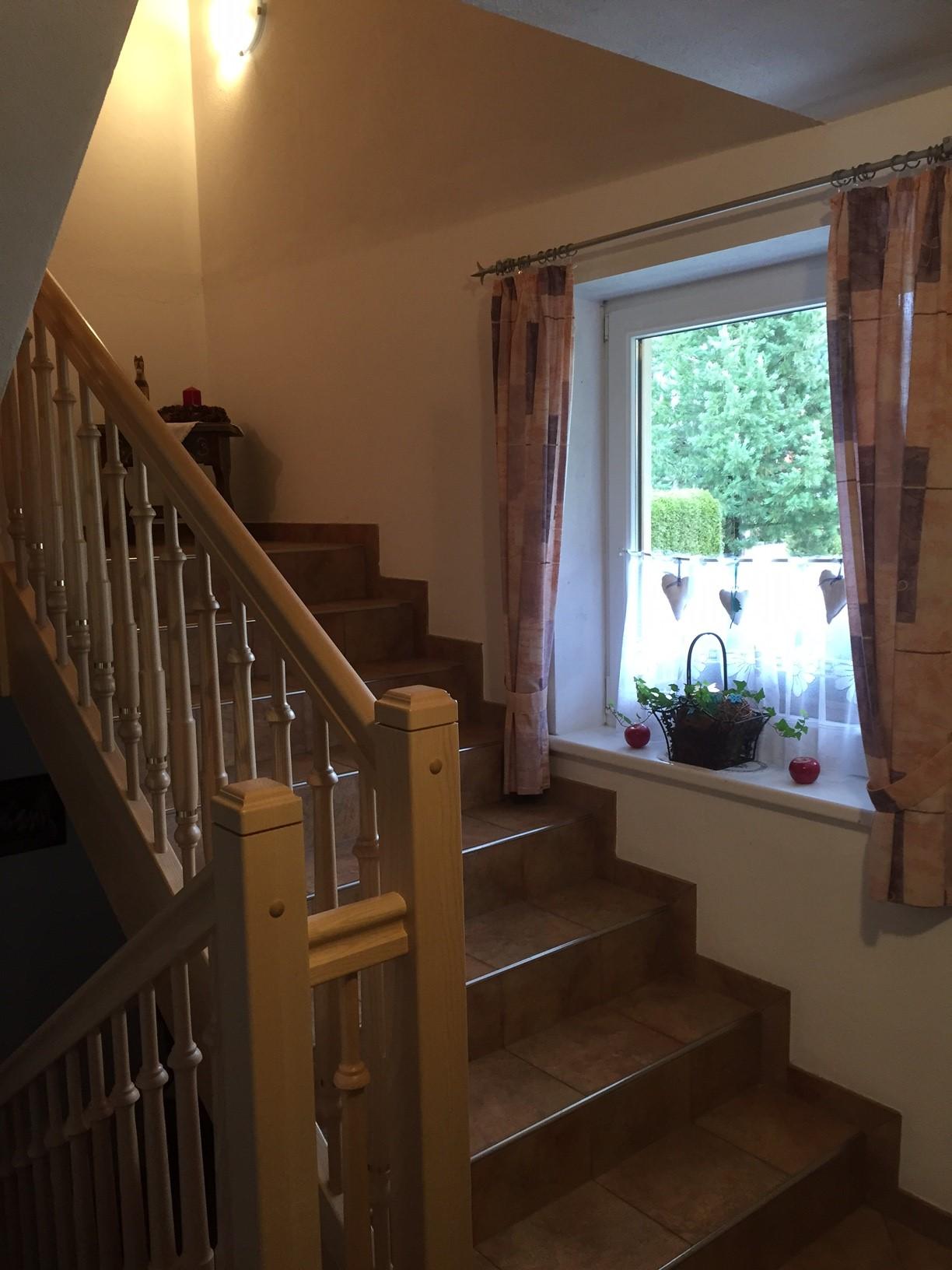 A bright staircase with a wooden staircase and a window through which the garden can be seen. The windows are decorated with curtains and there are plants on the windowsill.