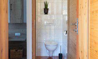 A modern bathroom with a bright, clean toilet. A green plant is on the wall and wooden elements are visible in the background.