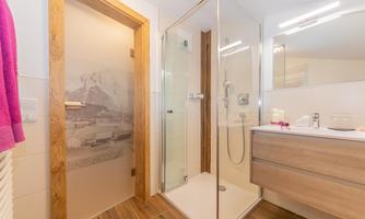 A modern bathroom with a glass shower and a wooden cabinet. The walls are light, and there is a decorative door with a mountain motif.