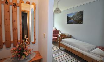 A cozy entrance area with a wooden panel and a mirror. In the background, there is a simple bedroom with a single bed and a picture frame on the wall.