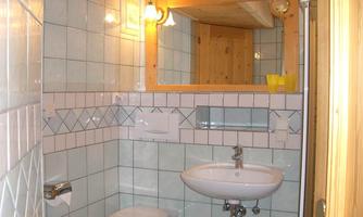 A simple bathroom with white tiles and a sink. In the background, a mirror and a wooden shelf are visible.