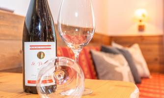 A bottle of wine is standing on a wooden table next to a wine glass. In the background, cozy cushions and warm light are visible.
