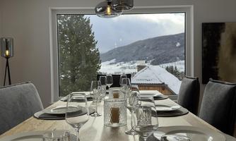 An elegant dining table in a bright room overlooking the snowy mountains. The table is beautifully set with dishes and glasses.