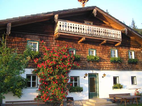 A traditional wooden house with a balcony and blooming plants. The facade is framed with colorful flowers and radiates a cozy atmosphere.