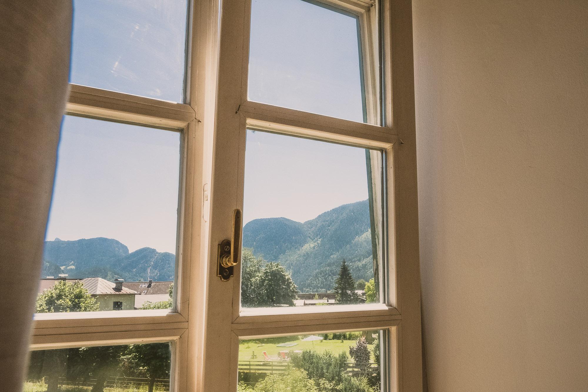 A window with a view of green meadows and snow-covered mountains. The sky is clear and blue.