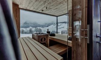 A modern room with wooden benches and a window front that offers a view of snow-covered mountains. The atmosphere is calm and relaxing.