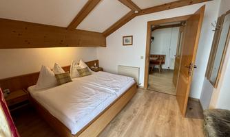 A cozy bedroom with two beds and a wooden ceiling. The door leads to an adjacent room with a table.