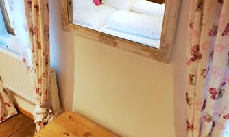 A cozy room with a wooden table and a mirror. In the mirror, the bed with white pillows can be seen.