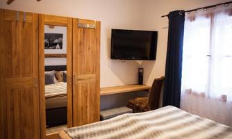 A cozy bedroom with a wooden wardrobe, a television, and a bed. The walls are light-colored and there are curtains at the windows.