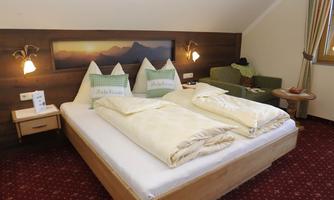 A cozy hotel room with a large double bed and two pillows. In the background, soft colors and a living room corner can be seen.