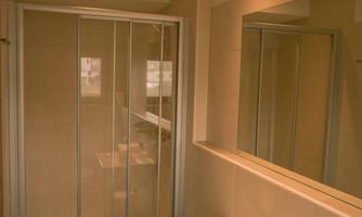 A modern bathroom with a shower and a sink. The walls are light tiled and there is a large mirror area.
