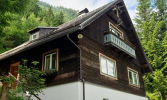 A charming wooden house in the mountains, surrounded by trees. The garden area is green and inviting.