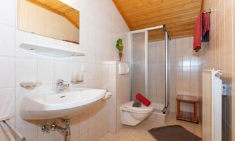 A modern bathroom with tiled walls and a shower. It features a sink, a toilet, and a wooden chair.