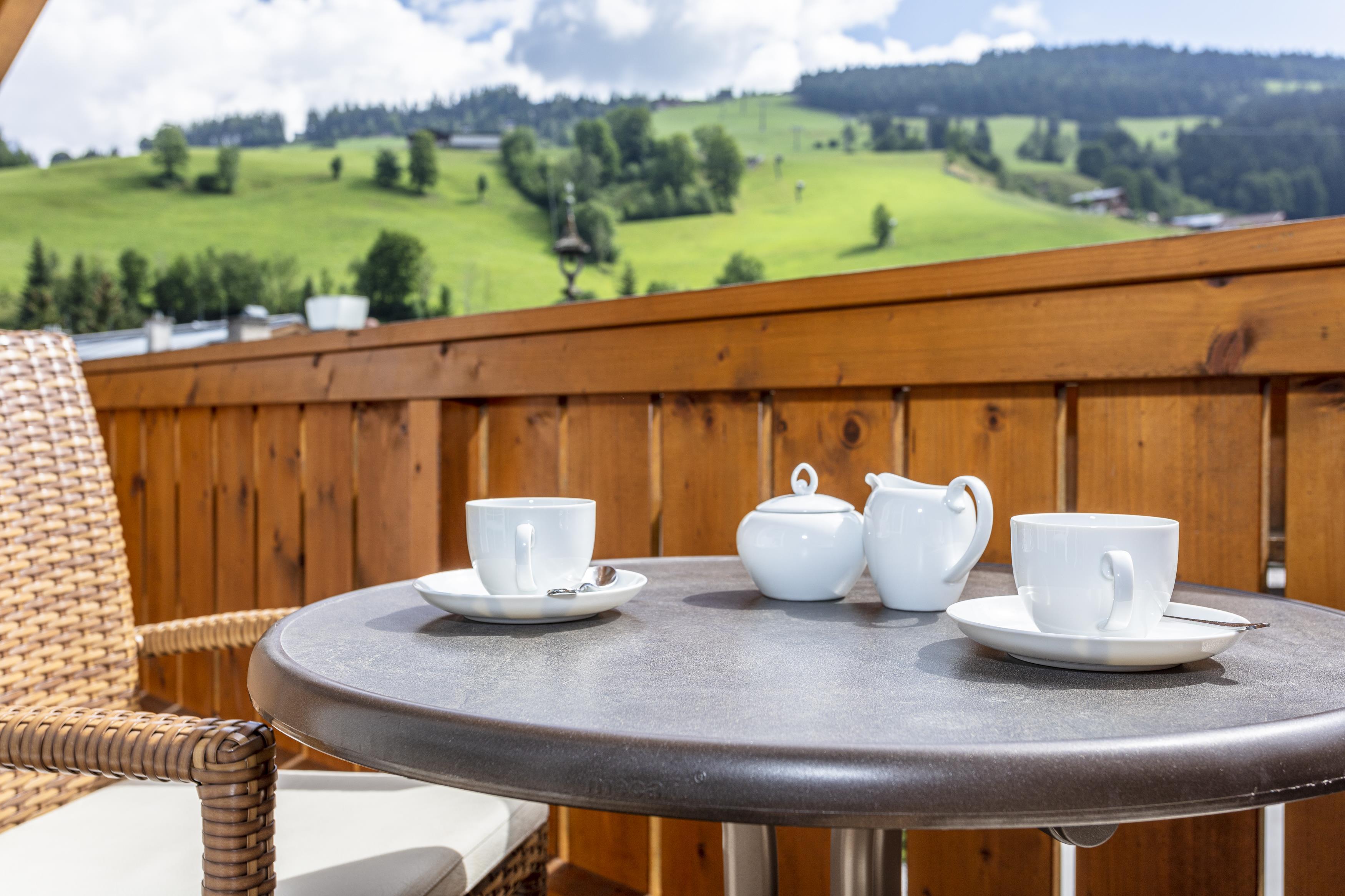 A beautiful balcony with a table on which two coffee cups and a teapot are placed. In the background, green hills and a cloudy but bright sky can be seen.