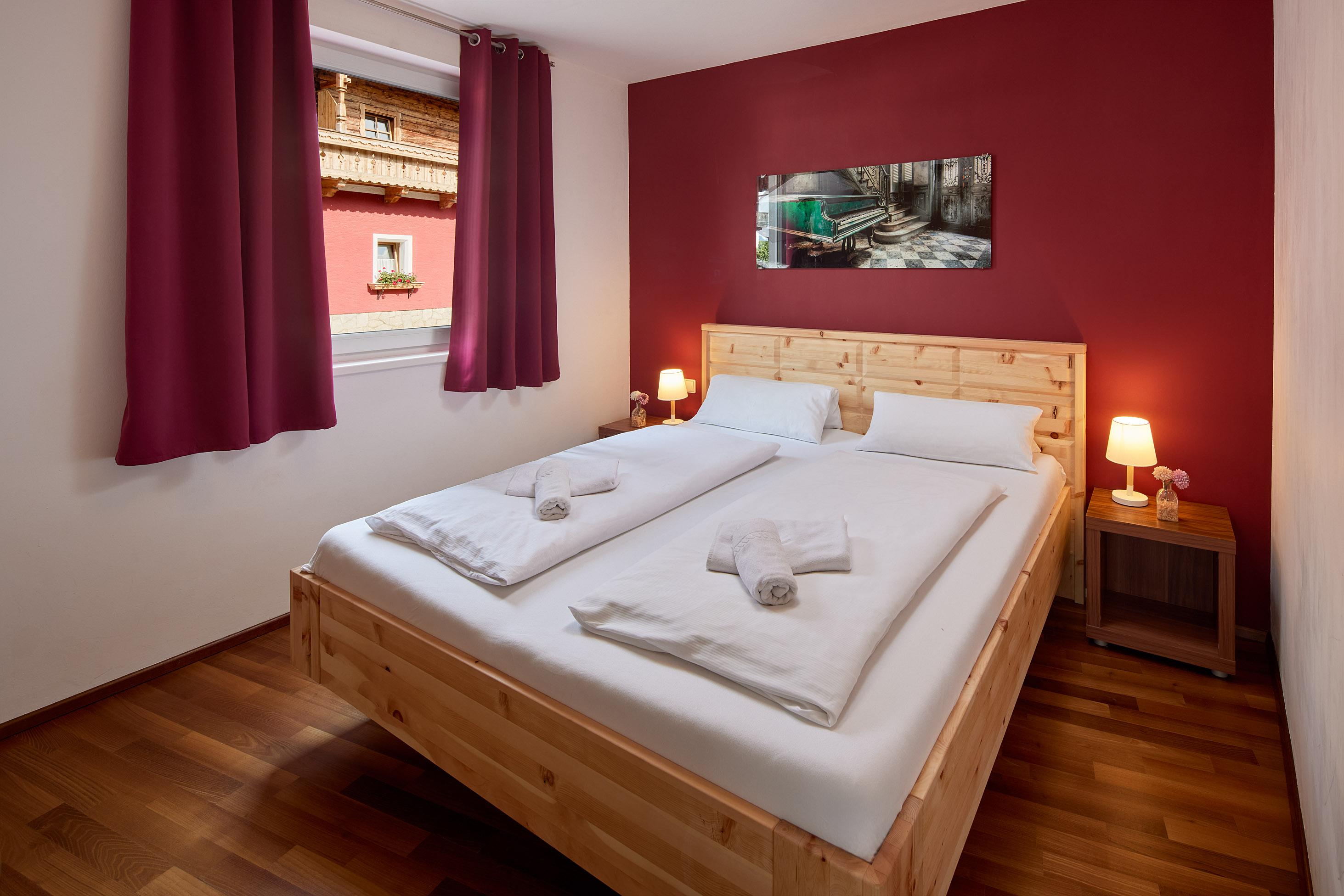 A cozy bedroom with a double bed and wooden flooring. The walls are painted red and there are windows with curtains.