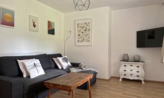 A cozy living space with a dark sofa and a wooden table. Decorative pictures hang on the walls, and a television is mounted.