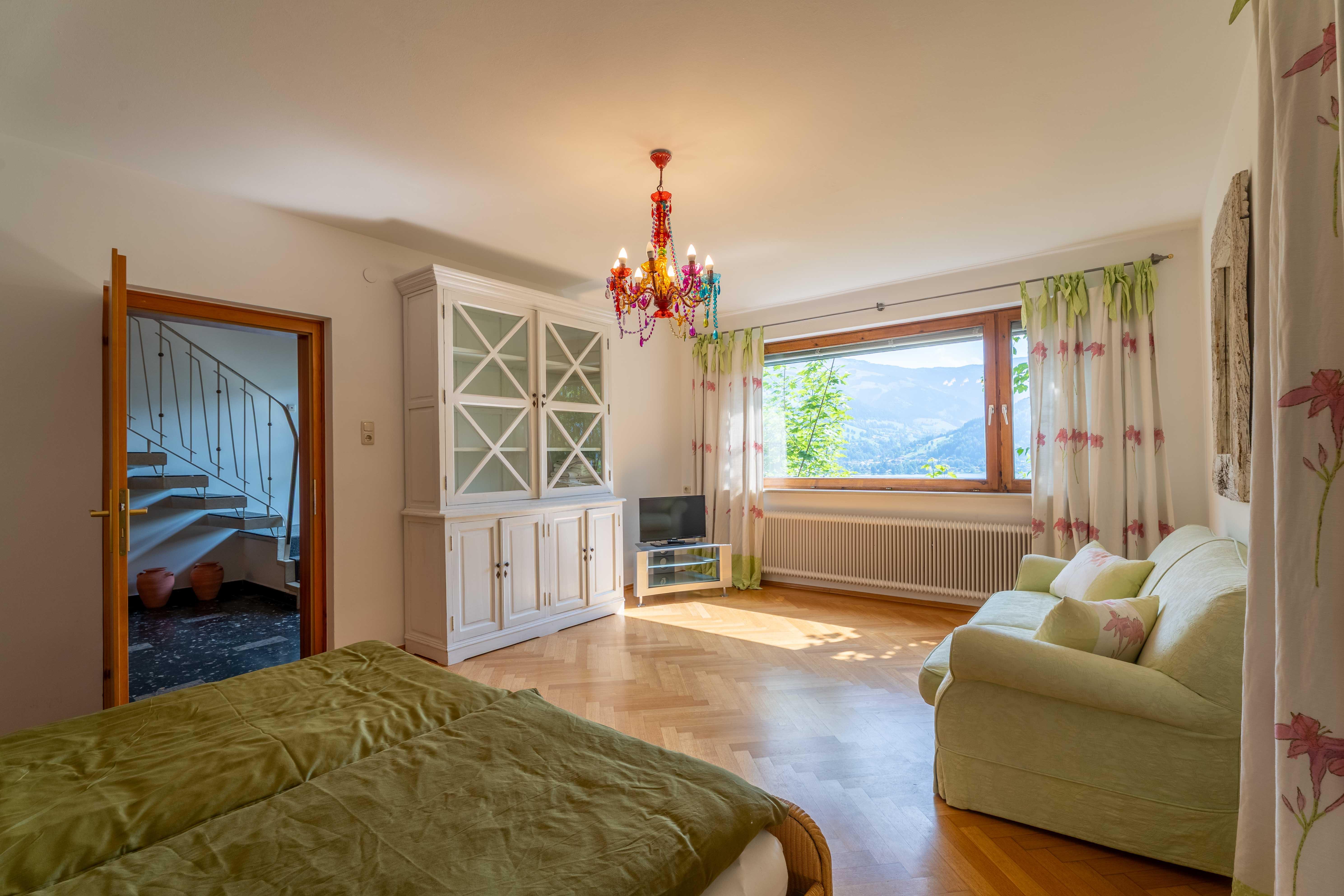 A bright bedroom with a green bed and a cozy sofa. Large windows provide a view of nature and let in plenty of light.