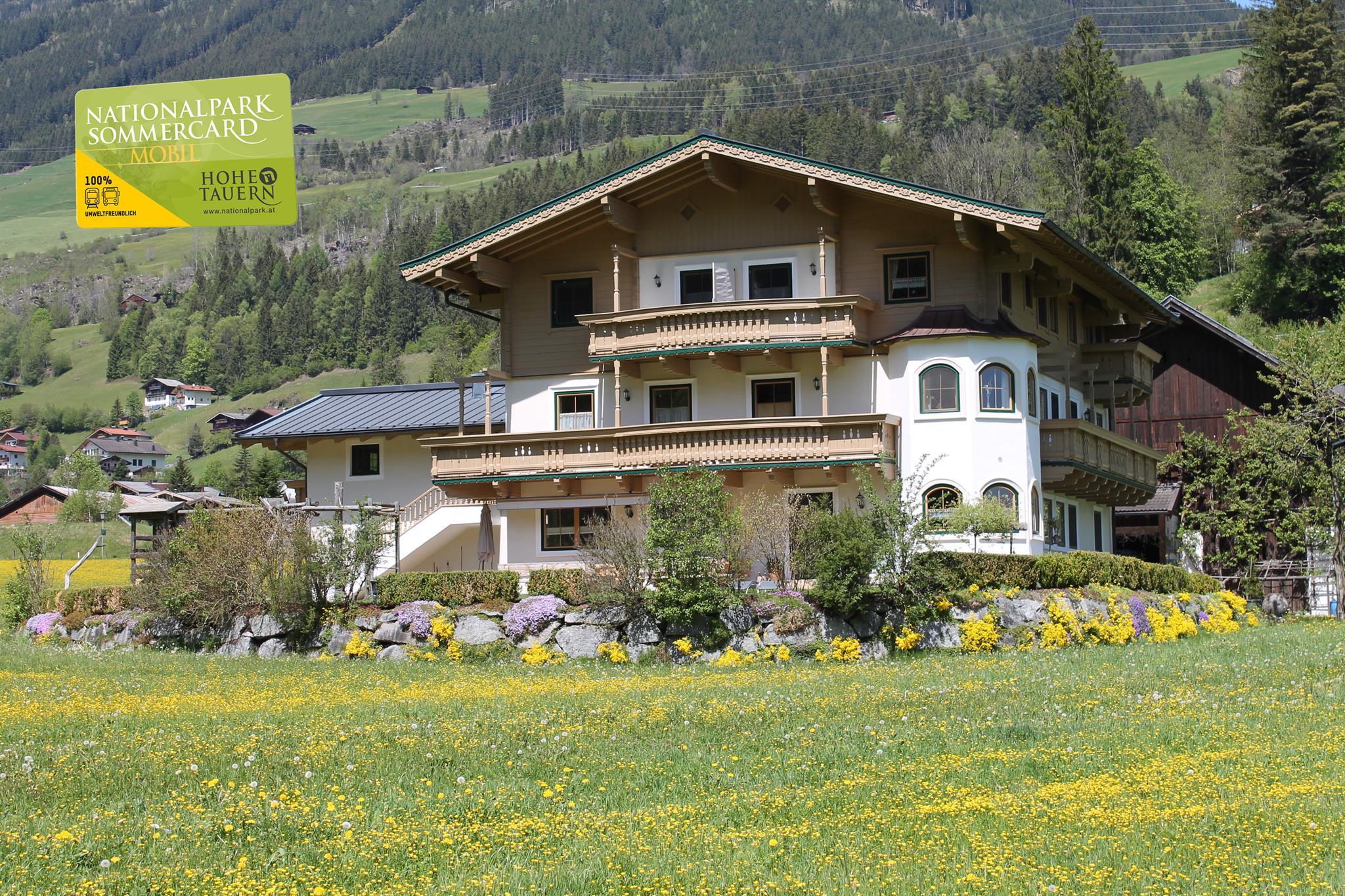 A typical Alpine house surrounded by meadows and mountains. The flowers bloom in vivid colors and the sky is clear and sunny.