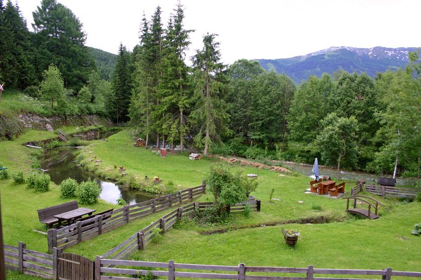 A green meadow with trees and a small stream. In the background, mountains and a cozy seating area can be seen.