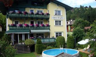 A charming yellow house with balconies full of flowers, surrounded by green trees. In the garden, there is a round swimming pool and a sunbathing area.