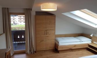 A bright room with wooden flooring, a window overlooking a balcony, and a single bed. The room is simply furnished with a wardrobe and a table.