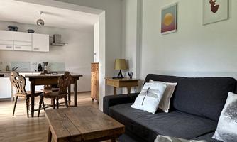 A cozy living room with a gray sofa and a wooden table. In the background, a bright kitchen with a small dining table is visible.