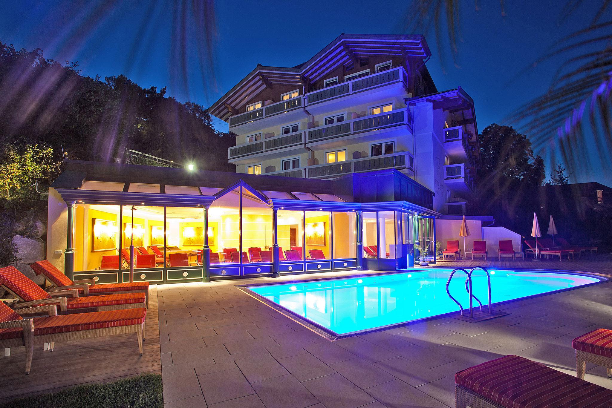 A modern hotel building with a illuminated pool area at night. The relaxing outdoor space features cozy loungers.
