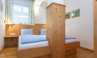 A bright room with two single beds and wooden furniture. Large windows provide plenty of natural light.
