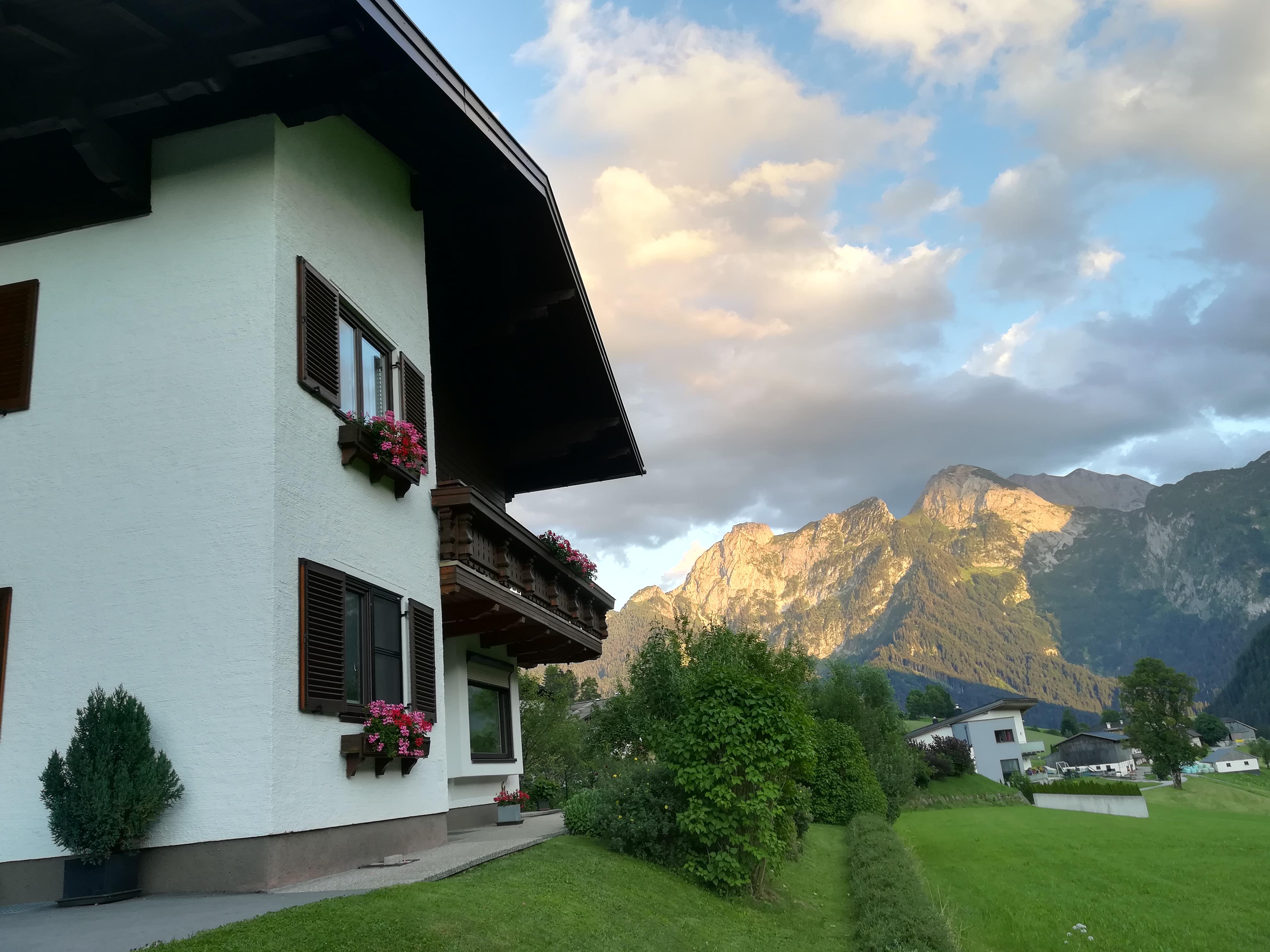 A charming house with balconies and flowers, surrounded by green meadows. In the background, impressive mountains rise under a cloudy sky.