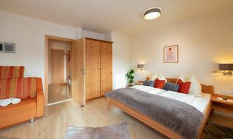 A cozy bedroom with a large bed and colorful pillows. There is also a small sitting area and a wooden wardrobe.