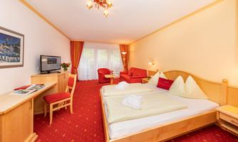 A cozy hotel room with a double bed, red chairs, and a desk. Large windows provide plenty of daylight.