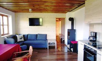 A cozy living room with a blue sofa, a television, and a modern kitchen. The space has a warm wooden ceiling and a wood stove.