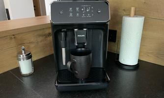 A modern coffee machine stands on a dark countertop. A cup is placed underneath, and there are various utensils in the background.