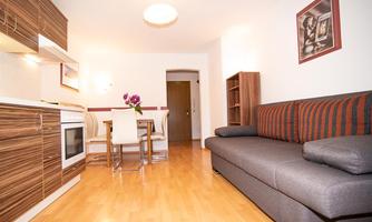 A modern apartment with an open kitchen and a cozy living area. The space is bright and well-furnished, featuring a dining table and a couch.