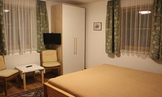 A simple room with a bed, two chairs, and a small table. Windows with curtains and a wardrobe are also visible.