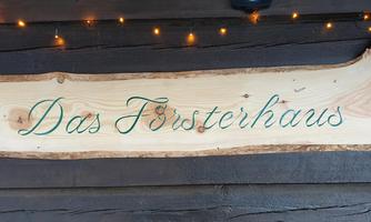 A wooden sign with the inscription "The Forester's House". It hangs on a dark wall and is decorated with small lights.